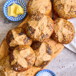 Whole Wheat Sourdough Pretzel Buns topped with different seeds on a wooden board, view from top. Framed by 2 little blue and white dishes with butter and flaky salt