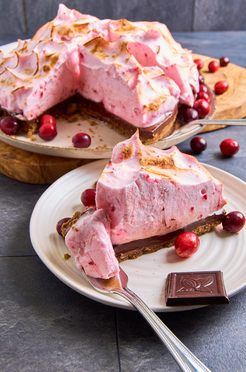 Cranberry Chocolate Meringue Pie with Pecan Crust now with caramelised peaks. View from front. A slice of it on a small plate, another waiting. Surrounded by chocloate pieces, pecans and cranberries.