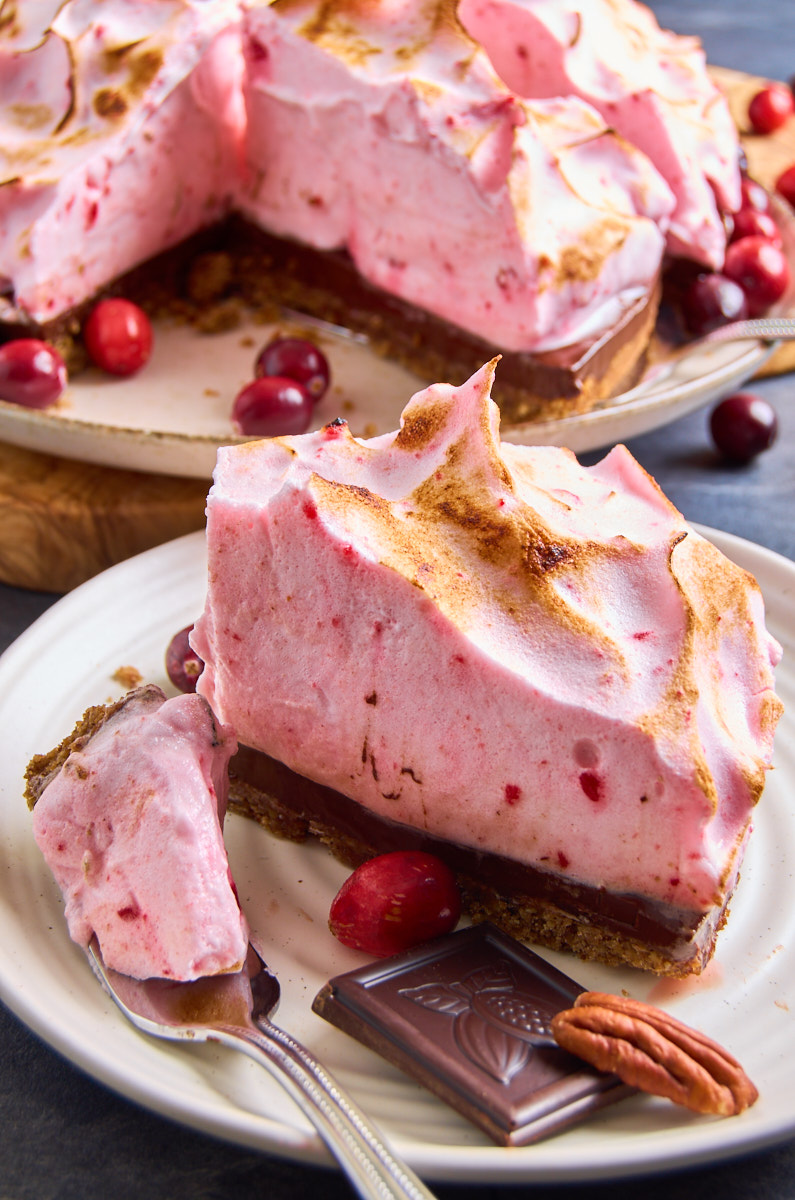 Cranberry Chocolate Meringue Pie with Pecan Crust now with caramelised peaks. View from front. A slice of it on a small plate, another waiting. Surrounded by chocloate pieces, pecans and cranberries.
