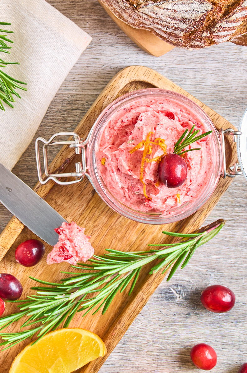 Whipped Honey Cranberry Butter With Orange and Rosemary in a little jar standing on a wooden board. Surrounded by fresh cranberries, orange slices and rosemary. A loaf of bread in the background.
