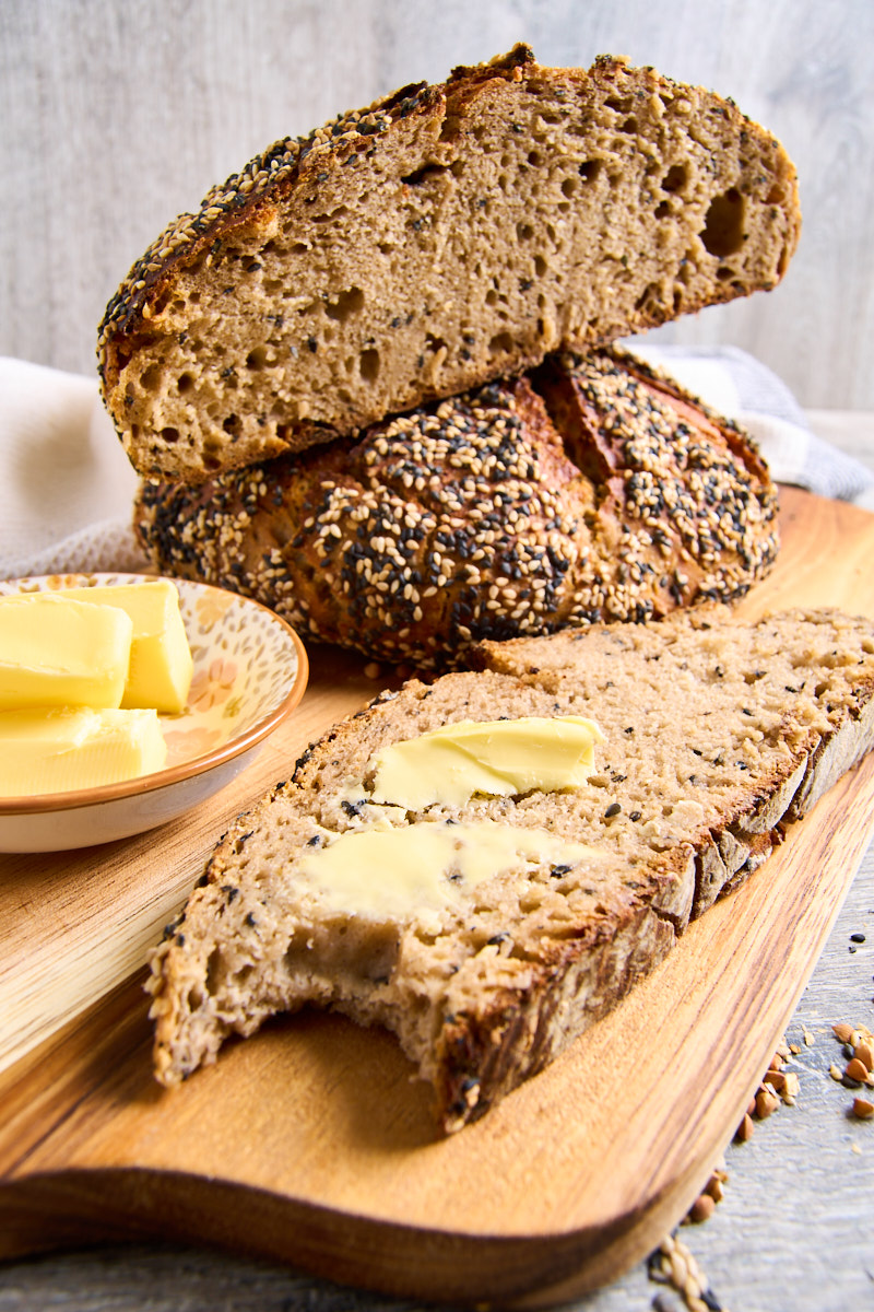 Sourdough Buckwheat No-Knead Bread cut in half, a slice with some butter on it in front, on a wooden board.