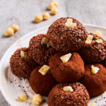 Chocolate Ginger Crunch Protein Balls stacked into a pyramid on a white plate, some rolled in cacao nibs, some in cacao powder, decorated with bits of crystallized ginger.